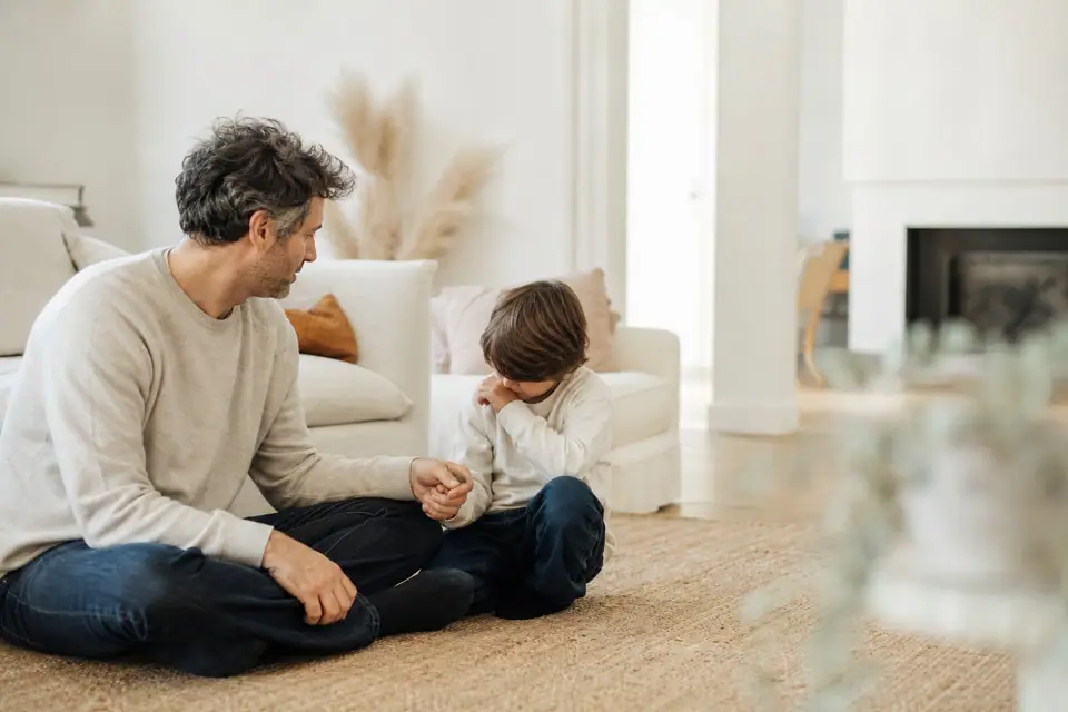 Parent assis au sol tenant calmement la main d’un jeune enfant, moment de réconfort dans un salon lumineux