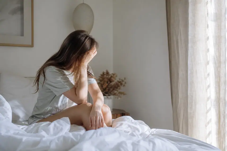Personne se tenant la tête, assise sur un lit un samedi matin, lumière douce à travers les rideaux