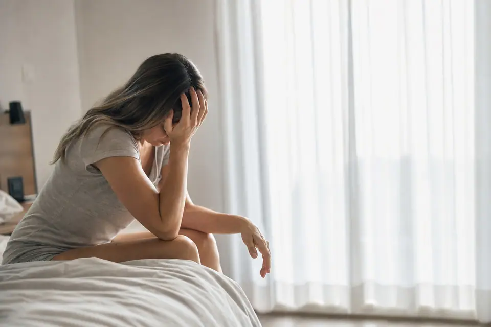 Personne assise au bord d’un lit, main sur le front, lumière douce du matin