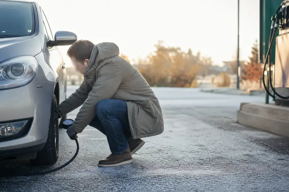 Automobiliste en parka accroupi près de la roue avant d’une voiture grise, dans une station-service par un matin froid