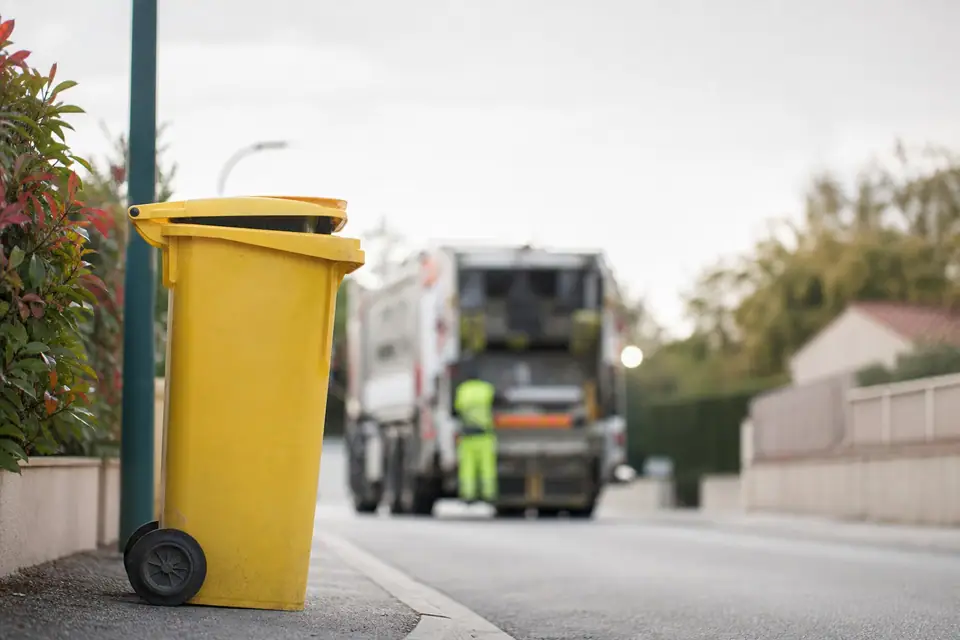 Bac de tri jaune posé au bord d’un trottoir résidentiel, couvercle entrouvert sur des emballages