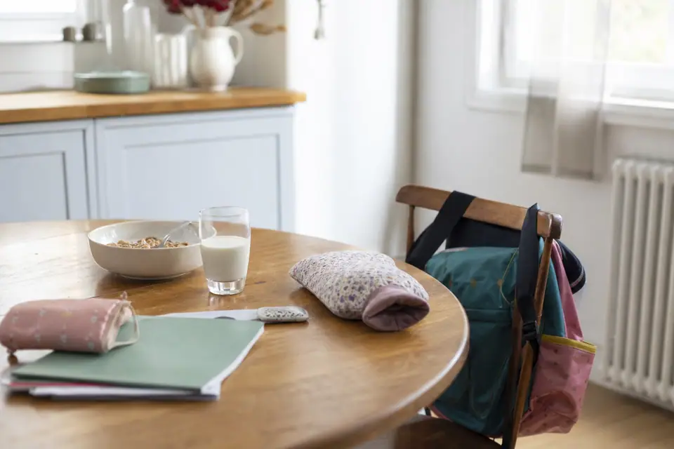 Table de petit-déjeuner avec bol de céréales à peine entamé, verre de lait, bouillotte et cartable sur une chaise